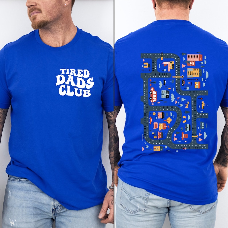 Puede incluir: Una camiseta azul real con el texto "TIRED DADS CLUB" en blanco en la parte delantera. La parte posterior de la camiseta presenta una ilustraci&oacute;n colorida de una ciudad con carreteras, edificios y coches.