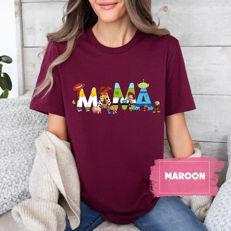 K&ouml;nnte beinhalten: Ein maroonfarbenes T-Shirt mit einem Aufdruck der Toy Story-Charaktere, die "MAMA" buchstabieren.