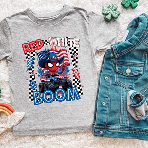 May include: A gray t-shirt featuring a graphic of a red Spider-Man character wearing a cowboy hat and driving a monster truck with the text "RED WHITE & BOOM". A denim jacket is next to the shirt.