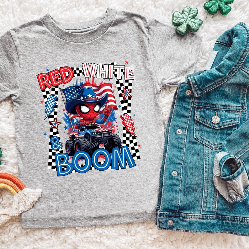 May include: A gray t-shirt featuring a graphic of a red Spider-Man character wearing a cowboy hat and driving a monster truck with the text "RED WHITE & BOOM". A denim jacket is next to the shirt.