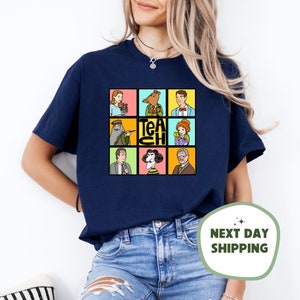 May include: Navy blue t-shirt featuring a colorful grid of cartoon characters and the word "TEACH". The design includes images of a woman with flowers, a rodent in a suit, and other figures. A fun and educational theme.
