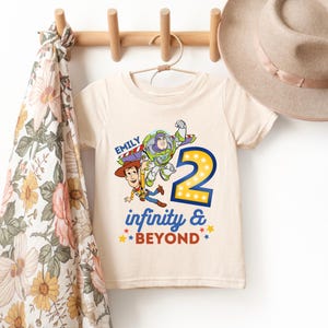 K&ouml;nnte beinhalten: Ein cremefarbenes T-Shirt mit einer Grafik von Buzz Lightyear und Woody aus Toy Story, mit der Zahl 2 in gelben Lichtern. Das Shirt hat auch den Text "Emily" und "infinity & BEYOND" in Blau und Rot.