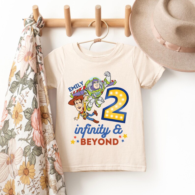 K&ouml;nnte beinhalten: Ein cremefarbenes T-Shirt mit einer Grafik von Buzz Lightyear und Woody aus Toy Story, mit der Zahl 2 in gelben Lichtern. Das Shirt hat auch den Text "Emily" und "infinity & BEYOND" in Blau und Rot.