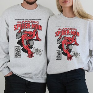May include: Two grey sweatshirts featuring a comic book style image of Spider-Man. The text on the shirts reads "Never has anyone fought such merciless foes as... the Amazing Spider-Man".