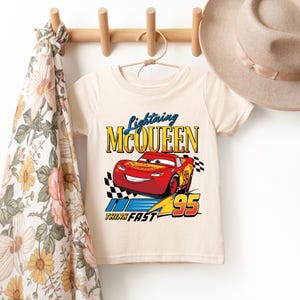 May include: Cream-colored short-sleeved t-shirt featuring a graphic of Lightning McQueen, a red race car with the number 95. The text "Lightning McQueen" is above the car, and "Think Fast" is below. A checkered flag design is on the right.