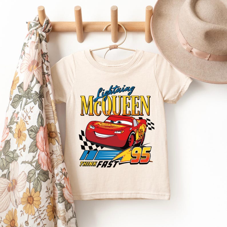 May include: Cream-colored short-sleeved t-shirt featuring a graphic of Lightning McQueen, a red race car with the number 95. The text "Lightning McQueen" is above the car, and "Think Fast" is below. A checkered flag design is on the right.