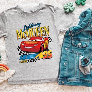 May include: A gray t-shirt featuring Lightning McQueen from the Cars movie. The design includes the text "Lightning McQueen" in blue and yellow, a red race car, and the number "95". A denim jacket is next to the shirt.