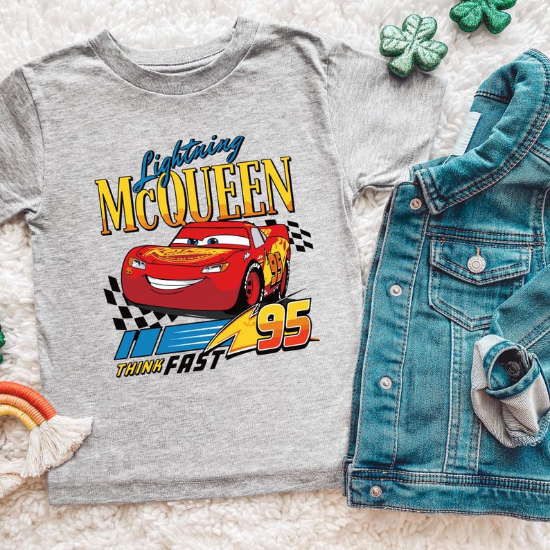 May include: A gray t-shirt featuring Lightning McQueen from the Cars movie. The design includes the text "Lightning McQueen" in blue and yellow, a red race car, and the number "95". A denim jacket is next to the shirt.