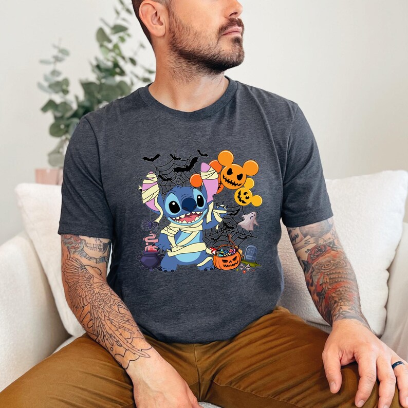 May include: Dark gray t-shirt featuring a Halloween-themed graphic. The design includes a blue cartoon character wrapped in bandages, surrounded by bats, spiderwebs, and jack-o'-lanterns. The character is holding a candy bucket. The shirt is a crew neck.