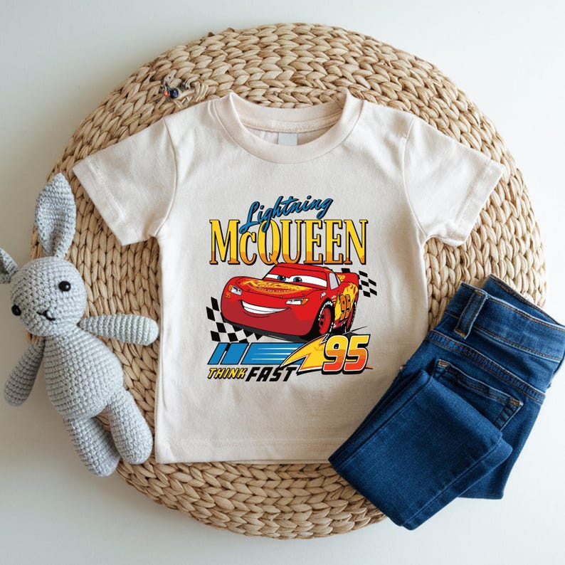 May include: A cream-colored t-shirt featuring a graphic of Lightning McQueen from the movie Cars. The design includes the text "Lightning McQueen", the number "95", and the phrase "Think Fast". Paired with blue denim jeans.