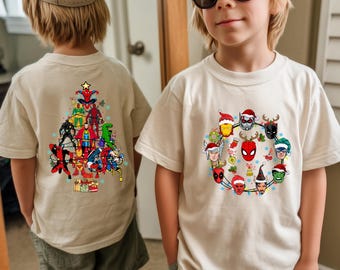 May include: Two beige t-shirts with superhero-themed Christmas designs. One shirt features a Christmas tree made of superheroes, the other has a wreath of superhero faces with Santa hats and festive decorations.