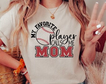May include: A beige t-shirt with the text "MY FAVORITE player CALLS ME MOM" in black and red. The design includes a baseball graphic. The shirt is a casual wear item.