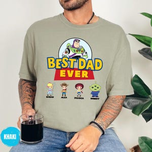 May include: Khaki t-shirt with a graphic of Buzz Lightyear and the text "BEST DAD EVER." Below the text are cartoon characters with names: Sarah, Alex, Jessica, and Robert. The shirt is worn by a person.