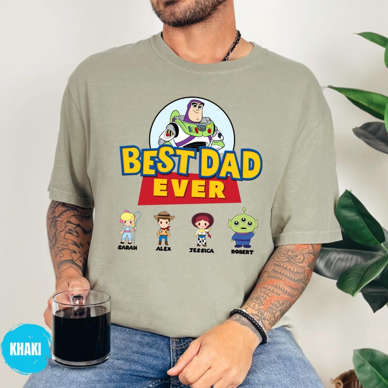 May include: Khaki t-shirt with a graphic of Buzz Lightyear and the text "BEST DAD EVER." Below the text are cartoon characters with names: Sarah, Alex, Jessica, and Robert. The shirt is worn by a person.