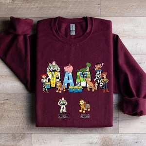 Może przedstawiać: Bordowa bluza z grafiką postaci z Toy Story, kt&oacute;re tworzą napis "MAMA" z nazwami "Noah" i "Jack" poniżej.