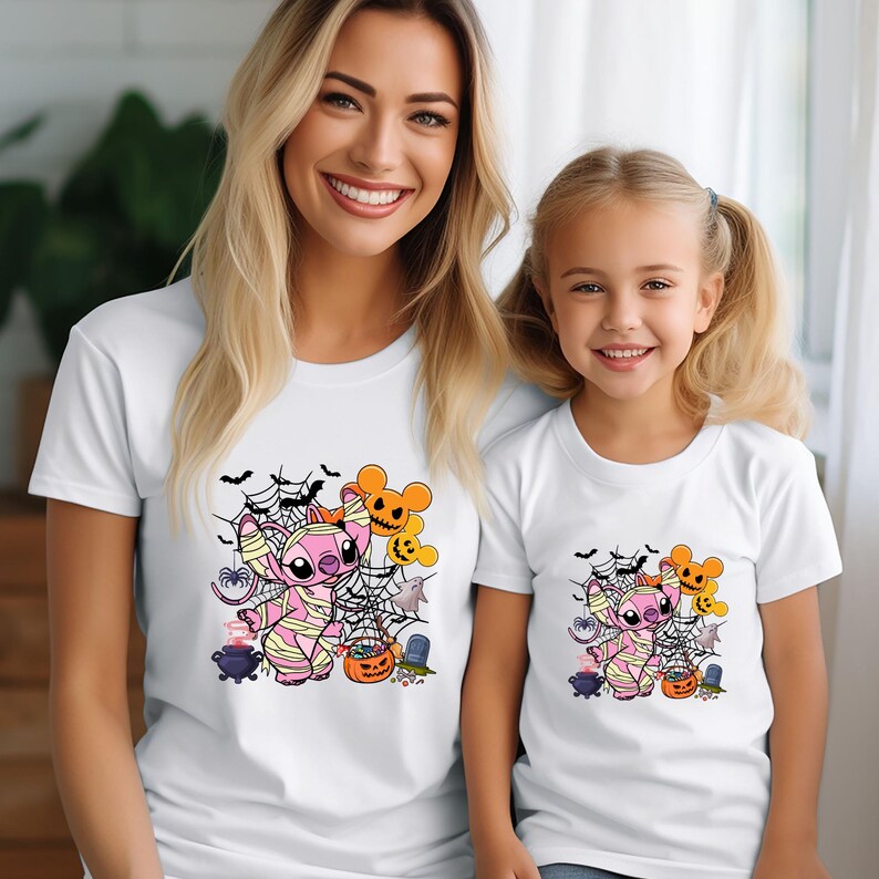 May include: White t-shirts featuring a Halloween-themed graphic. The design includes a pink cartoon character wrapped in bandages, spiderwebs, bats, pumpkins, and other Halloween elements. The shirts are for adults and children.
