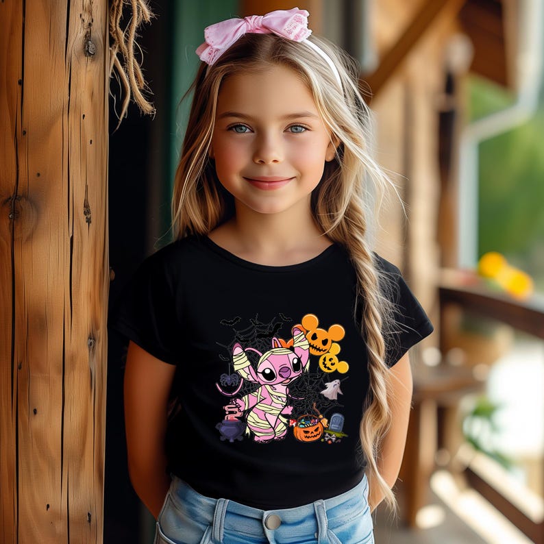 May include: A black t-shirt featuring a Halloween-themed graphic. The graphic includes a pink character wrapped in bandages, a jack-o'-lantern, and other spooky elements. The shirt is worn by a young girl with blonde hair and a pink headband.