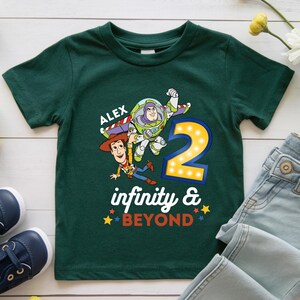 K&ouml;nnte beinhalten: Ein dunkelgr&uuml;nes T-Shirt mit einer Grafik von Buzz Lightyear und Woody aus Toy Story. Die Zahl "2" wird mit den Worten "infinity & BEYOND" angezeigt. Der Name "ALEX" ist &uuml;ber den Figuren aufgedruckt.
