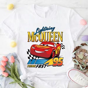 May include: White t-shirt featuring a graphic of Lightning McQueen from the movie Cars. The design includes the text "Lightning McQueen" in yellow and blue, the number "95", and the phrase "Think Fast".