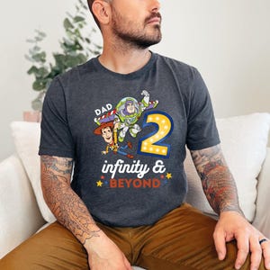 K&ouml;nnte beinhalten: Dunkelgraues T-Shirt mit einer farbenfrohen Grafik von Woody, Buzz Lightyear und der Zahl 2 mit dem Text "Dad infinity & beyond". Das Design enth&auml;lt gelbe, rote, blaue und wei&szlig;e Akzente. Das Shirt ist aus weichem Material.