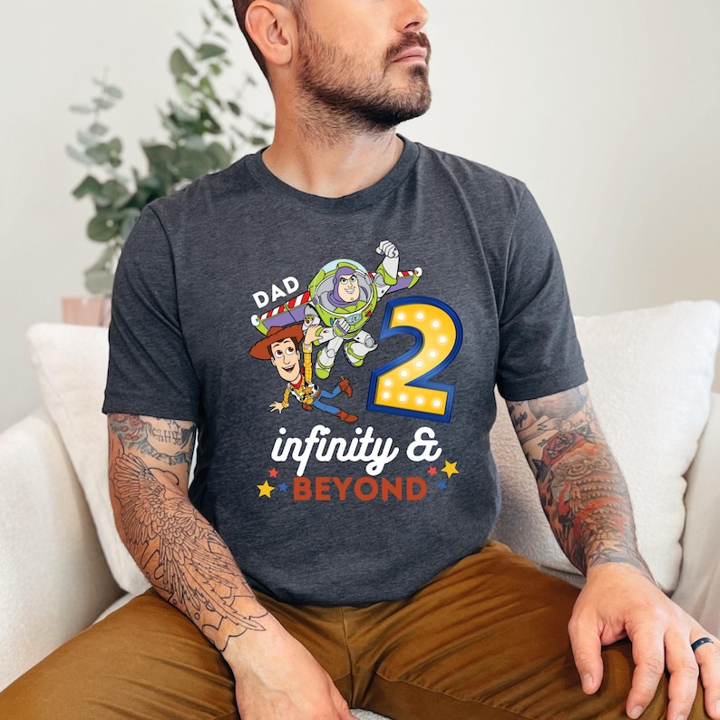 K&ouml;nnte beinhalten: Dunkelgraues T-Shirt mit einer farbenfrohen Grafik von Woody, Buzz Lightyear und der Zahl 2 mit dem Text "Dad infinity & beyond". Das Design enth&auml;lt gelbe, rote, blaue und wei&szlig;e Akzente. Das Shirt ist aus weichem Material.