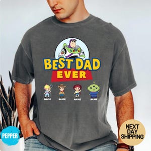 May include: Dark grey t-shirt with the text "BEST DAD EVER" in yellow and red, and a Buzz Lightyear graphic. Below are cartoon characters with the word "NAME" under each. The t-shirt is a casual style.