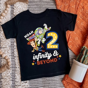 K&ouml;nnte beinhalten: Schwarzes Kleinkind-T-Shirt mit Buzz Lightyear und Woody aus Toy Story. Das Shirt hat die Zahl "2" mit Lichtern, den Namen "Noah" und den Satz "infinity & beyond" in farbigem Text.