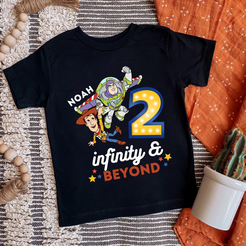 K&ouml;nnte beinhalten: Schwarzes Kleinkind-T-Shirt mit Buzz Lightyear und Woody aus Toy Story. Das Shirt hat die Zahl "2" mit Lichtern, den Namen "Noah" und den Satz "infinity & beyond" in farbigem Text.
