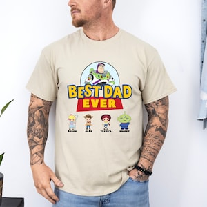 May include: Beige t-shirt featuring the phrase "BEST DAD EVER" in yellow and blue, with a red banner. The design includes a cartoon image of Buzz Lightyear and four character illustrations with names below. A casual tee.
