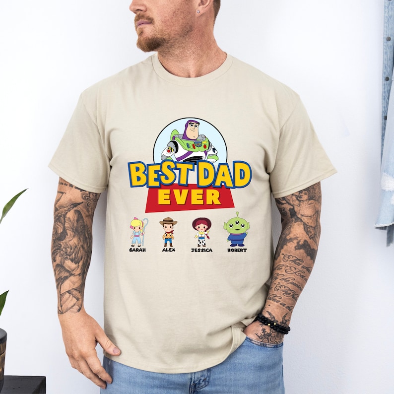 May include: Beige t-shirt featuring the phrase "BEST DAD EVER" in yellow and blue, with a red banner. The design includes a cartoon image of Buzz Lightyear and four character illustrations with names below. A casual tee.