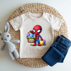 Puede incluir: Una camiseta color crema con un personaje de dibujos animados de Spider-Man sosteniendo una cesta de huevos de Pascua. La cesta tiene las palabras "Happy Easter". Unos vaqueros azules y un conejito de peluche gris tambi&eacute;n est&aacute;n en la imagen.