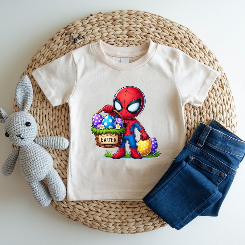 Puede incluir: Una camiseta color crema con un personaje de dibujos animados de Spider-Man sosteniendo una cesta de huevos de Pascua. La cesta tiene las palabras "Happy Easter". Unos vaqueros azules y un conejito de peluche gris tambi&eacute;n est&aacute;n en la imagen.