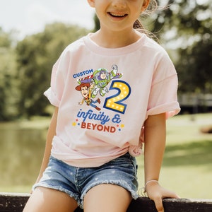 May include: Pink t-shirt with a graphic of Buzz Lightyear and Woody from Toy Story. The graphic includes the number 2 with lights around it and the text "infinity & BEYOND".