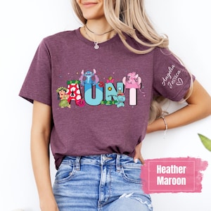 Puede incluir: Una camiseta de color marr&oacute;n heather con un gr&aacute;fico de la palabra "AUNT" deletreada con personajes de Stitch de la pel&iacute;cula de Disney Lilo & Stitch. La camiseta tiene los nombres "Angelia Jessica" escritos en la manga.