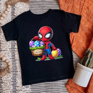 Puede incluir: Camiseta negra de manga corta con un dise&ntilde;o de Spider-Man de dibujos animados sosteniendo una cesta de huevos de Pascua. La cesta tiene las palabras "Happy Easter". Los huevos est&aacute;n decorados con varios patrones y colores.