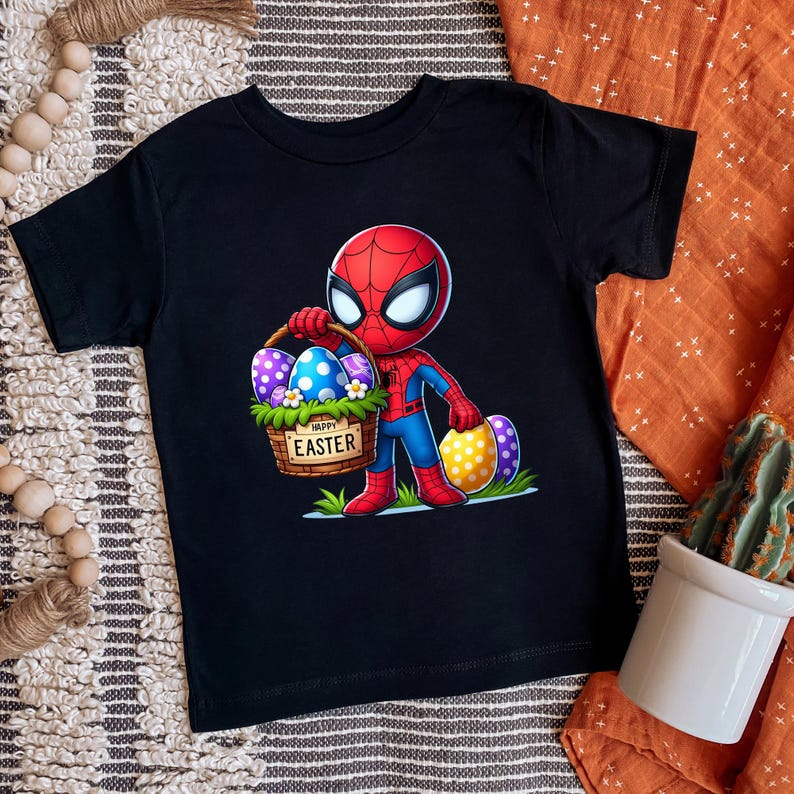 Puede incluir: Camiseta negra de manga corta con un dise&ntilde;o de Spider-Man de dibujos animados sosteniendo una cesta de huevos de Pascua. La cesta tiene las palabras "Happy Easter". Los huevos est&aacute;n decorados con varios patrones y colores.