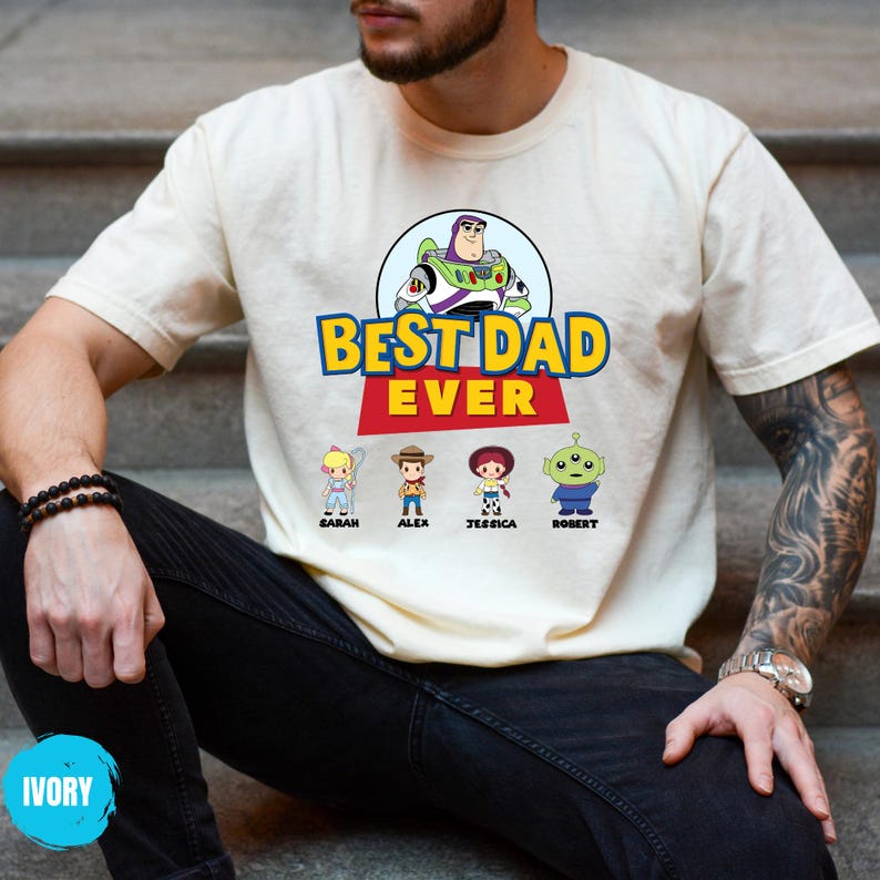 May include: Ivory t-shirt with a graphic of Buzz Lightyear and the text "BEST DAD EVER." Below the text are cartoon characters labelled Sarah, Alex, Jessica, and Robert. The shirt is being worn by a person.