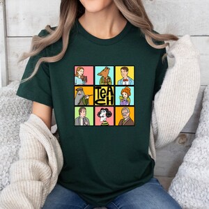 May include: Dark green t-shirt featuring a colorful graphic with nine cartoon portraits and the word "TEACH". The portraits include a woman with flowers, a bear in a suit, a man in a suit, a man with a beard, a woman with a pointer, a woman with flowers, a man in a suit, a woman with a black and white hairstyle, and an older man.