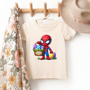 Puede incluir: Una camiseta color crema con un dibujo animado de Spider-Man sosteniendo una cesta de huevos de Pascua. La cesta tiene las palabras "Happy Easter". Dos huevos de Pascua adicionales est&aacute;n al lado de Spider-Man. Un sombrero y una bufanda est&aacute;n en el fondo.