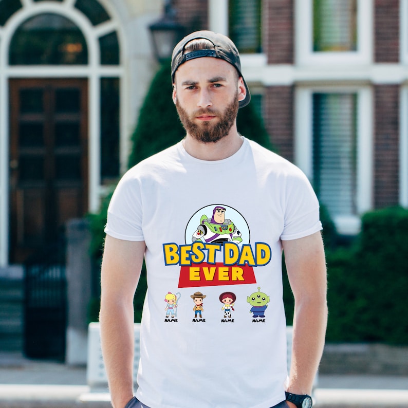 May include: White t-shirt with a colorful graphic featuring Buzz Lightyear and the text "BEST DAD EVER." Below the text are cartoon characters. The shirt is worn by a person wearing a black baseball cap.