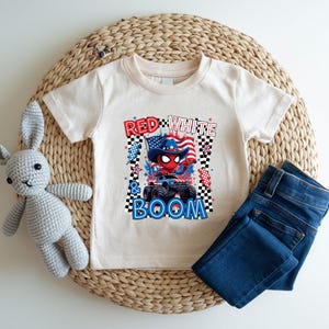 May include: A cream-colored t-shirt featuring a graphic with the text "Red, White & Boom." The graphic includes a monster truck, an American flag, and a character wearing a patriotic hat. Dark blue jeans and a stuffed bunny are also in the image.