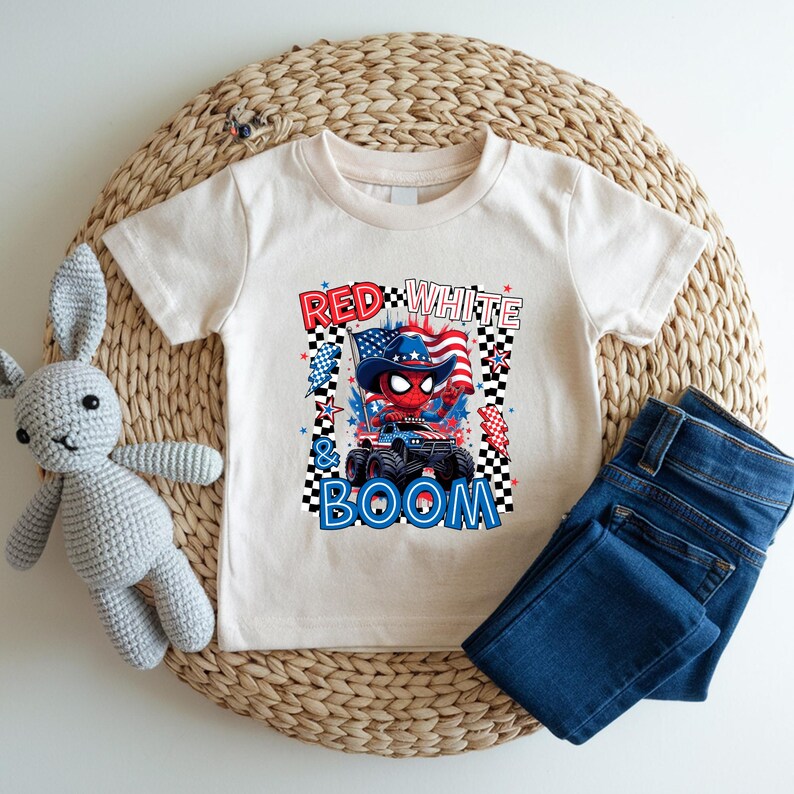 May include: A cream-colored t-shirt featuring a graphic with the text "Red, White & Boom." The graphic includes a monster truck, an American flag, and a character wearing a patriotic hat. Dark blue jeans and a stuffed bunny are also in the image.