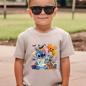May include: A beige short-sleeve t-shirt featuring a Halloween-themed graphic. The graphic includes a blue cartoon character wrapped in bandages, surrounded by bats, spiderwebs, and jack-o'-lanterns. The shirt is worn by a child wearing black sunglasses.