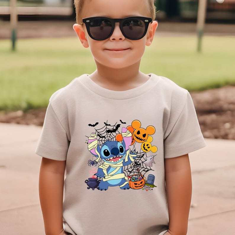 May include: A beige short-sleeve t-shirt featuring a Halloween-themed graphic. The graphic includes a blue cartoon character wrapped in bandages, surrounded by bats, spiderwebs, and jack-o'-lanterns. The shirt is worn by a child wearing black sunglasses.