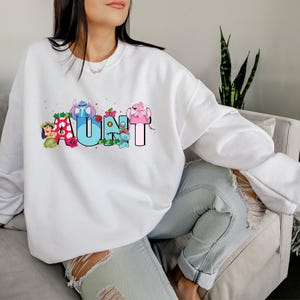 May include: White sweatshirt with a graphic design of the word "AUNT" spelled out with cartoon characters. The characters are from the Disney movie Lilo & Stitch.