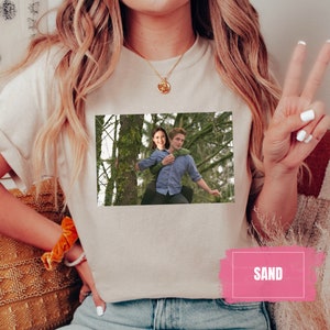 May include: A white t-shirt with a printed image of a couple in a forest. The image shows a woman with long brown hair and a man with short brown hair. The woman is wearing a green shirt and the man is wearing a blue shirt. The text "SAND" is printed in pink on the bottom right corner of the image.