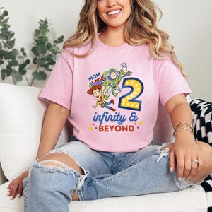 K&ouml;nnte beinhalten: Hellrosa T-Shirt mit einem farbenfrohen Aufdruck von Woody, Buzz Lightyear und der Zahl 2, mit den Worten "infinity & beyond". Das Wort "Mom" steht &uuml;ber der Grafik. Das T-Shirt wird von einer Person getragen.