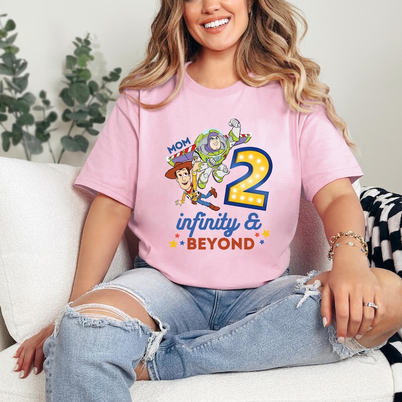 K&ouml;nnte beinhalten: Hellrosa T-Shirt mit einem farbenfrohen Aufdruck von Woody, Buzz Lightyear und der Zahl 2, mit den Worten "infinity & beyond". Das Wort "Mom" steht &uuml;ber der Grafik. Das T-Shirt wird von einer Person getragen.