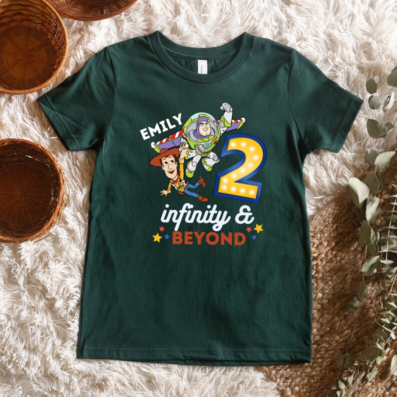 May include: Green t-shirt with a graphic of Woody and Buzz Lightyear from Toy Story. The shirt says "Emily" and "infinity & BEYOND" with a number 2 in the center.