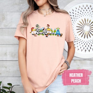 K&ouml;nnte beinhalten: Ein pfirsichfarbenes T-Shirt mit einer Grafik der Toy Story-Charaktere, die "Oma" buchstabieren.